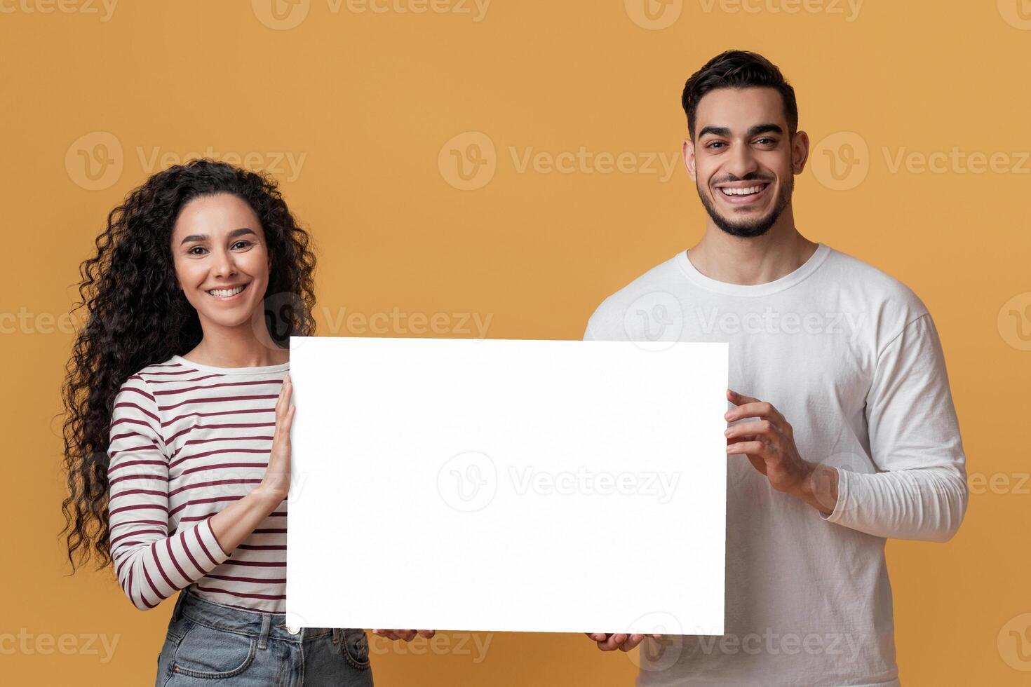 Ad Mockup. Happy Young Arab Spouses Holding White Blank Placard In Hands, Smiling Middle Eastern Couple Demonstrating Copy Space For Your Design While Standing Over Yellow Studio Background photo