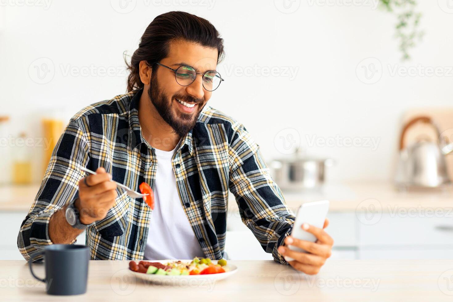 Handsome young arab long-haired, bearded man using cellphone while having healthy and tasty breakfast, checking social media or watching funny videos online, sitting at kitchen alone, copy space photo
