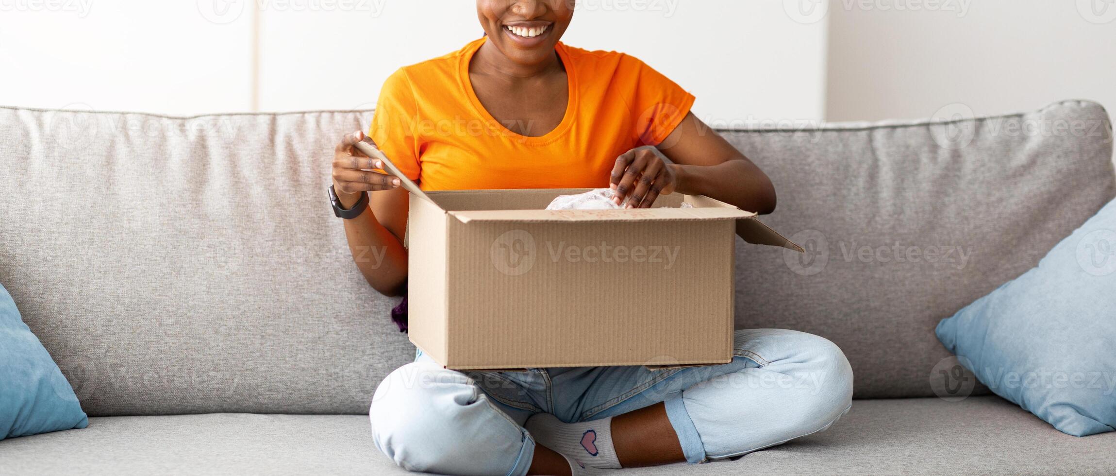 Full length of positive black lady opening cardboard package, receiving online store delivery at home. Young female client unboxing web shop order, satisfied with customer service photo