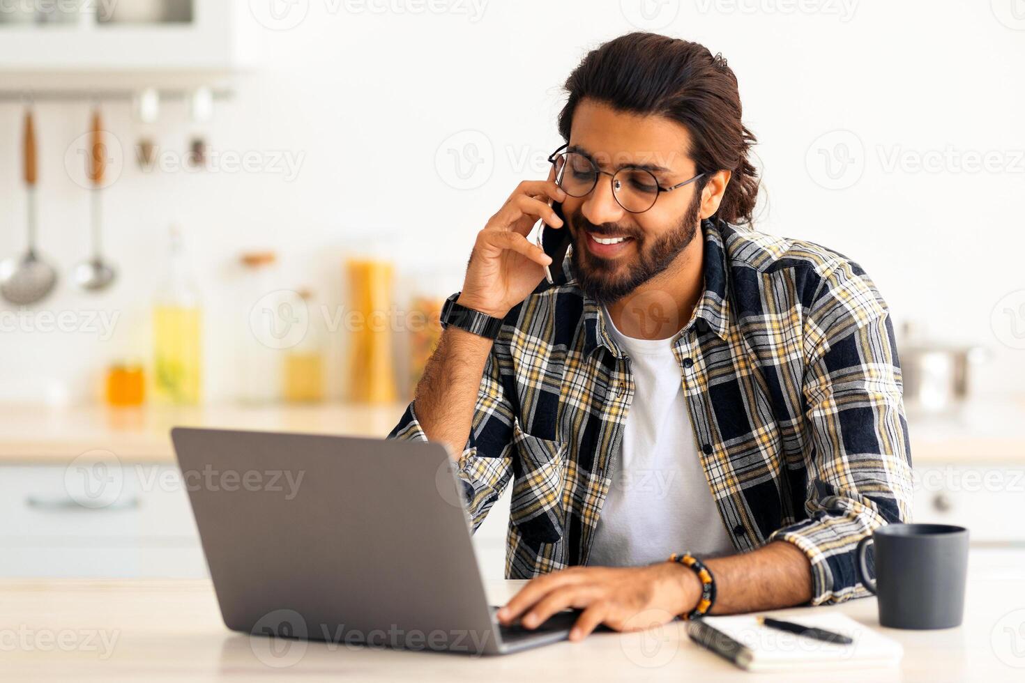 Busy young arab man freelancer talking on mobile phone and looking at laptop screen, working from home during isolation, copy space. Happy guy having conversation on phone at kitchen photo