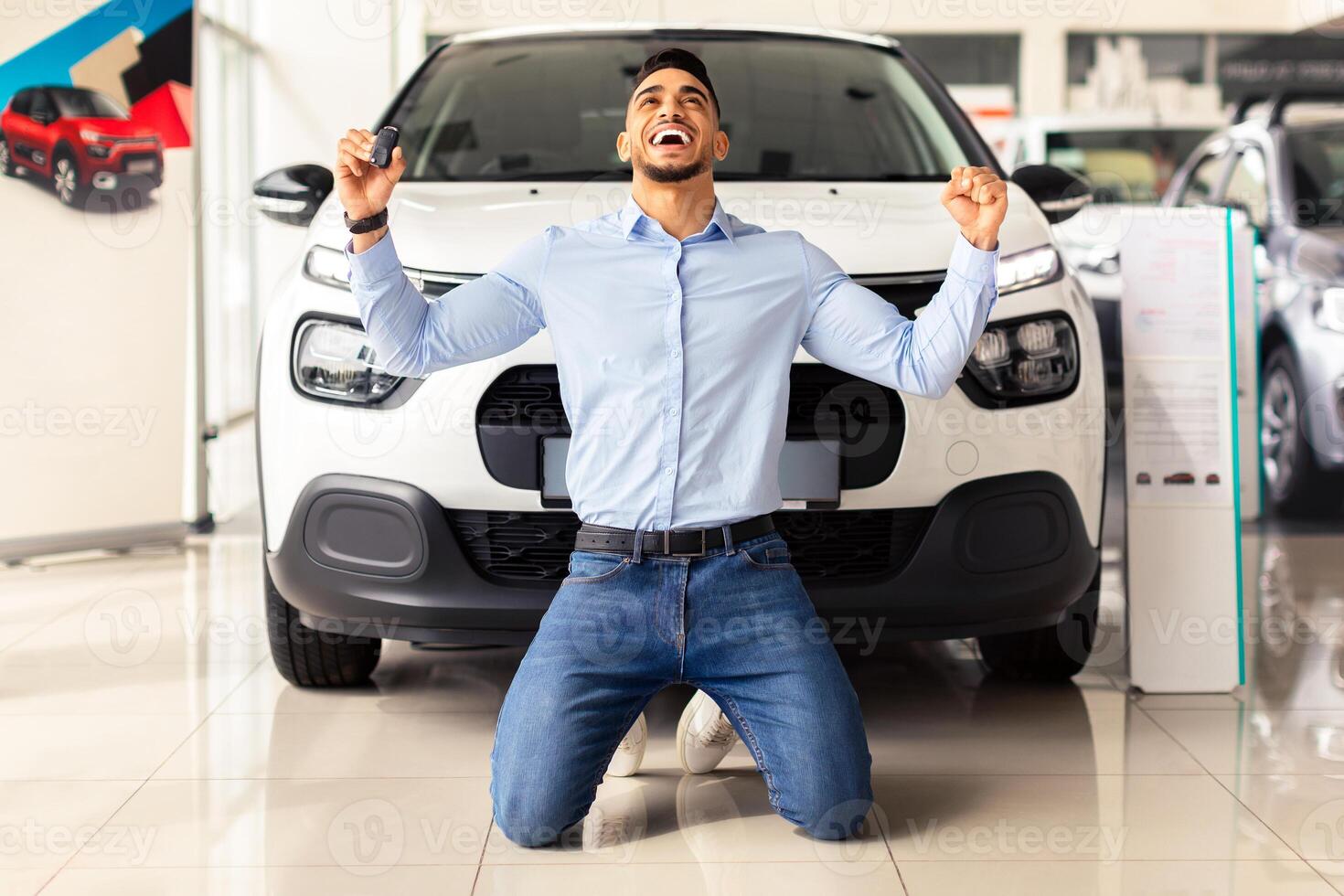 Emotional arab guy customer kneeling next to luxury white car in showroom, clenching fists and looking up, happy middle-eastern young man purchasing brand new auto of his dream, car retail concept photo