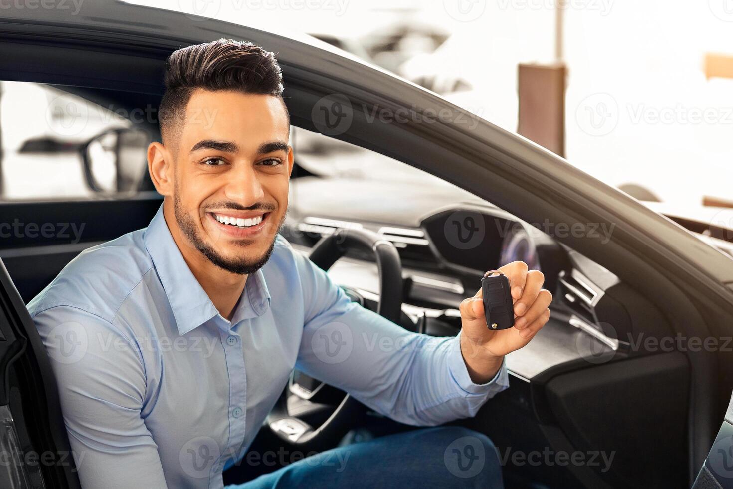 Handsome arab man customer sitting in auto and holding key, smiling at camera, buying car in showroom. Cheerful middle-eastern guy sitting inside nice sports car, happy to get new automobile photo