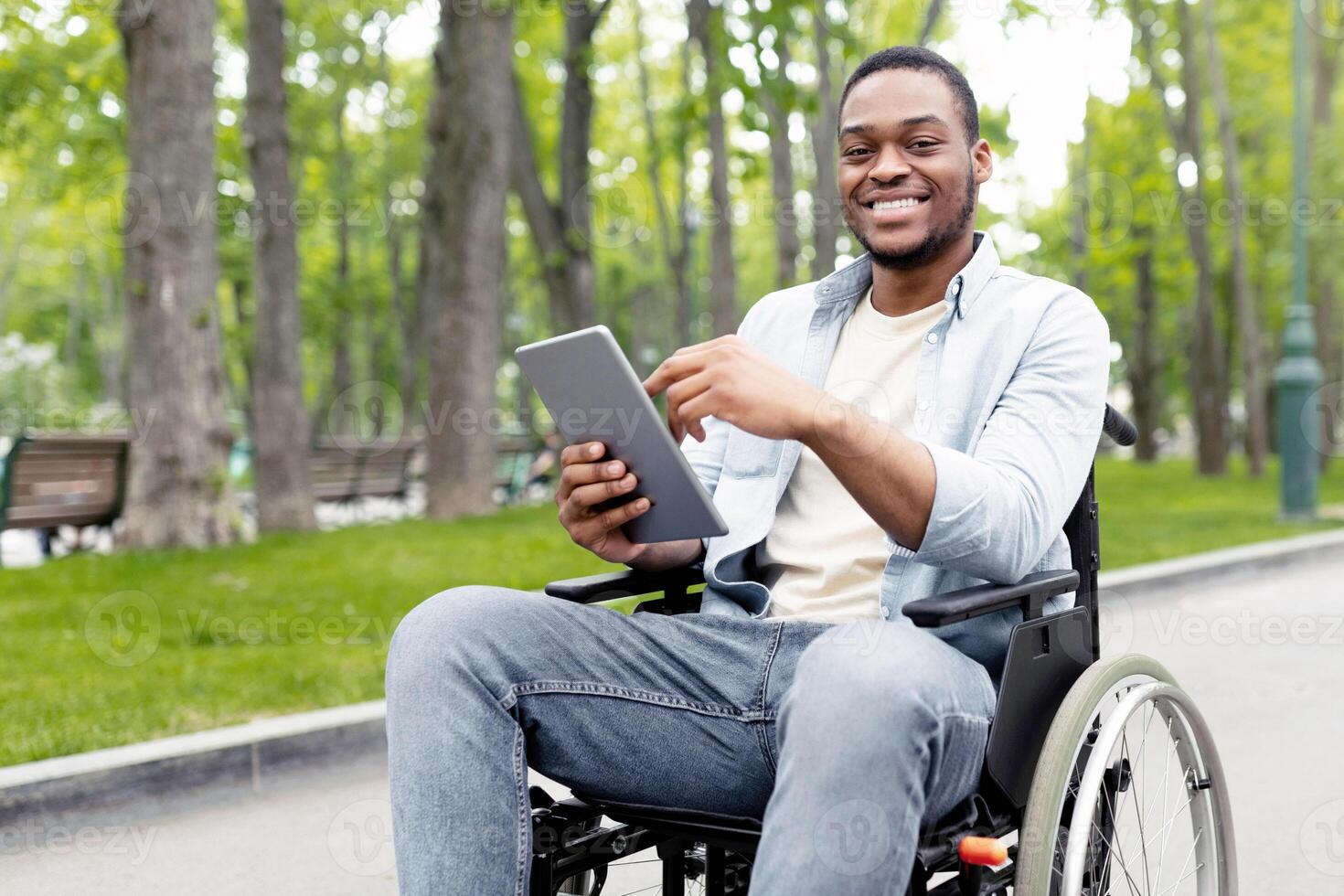 Portrait of cheeful impaired young man in wheelchair using laptop for remote communication at city park. Happy black guy with disability having online meeting, chatting with friend on web outdoors photo
