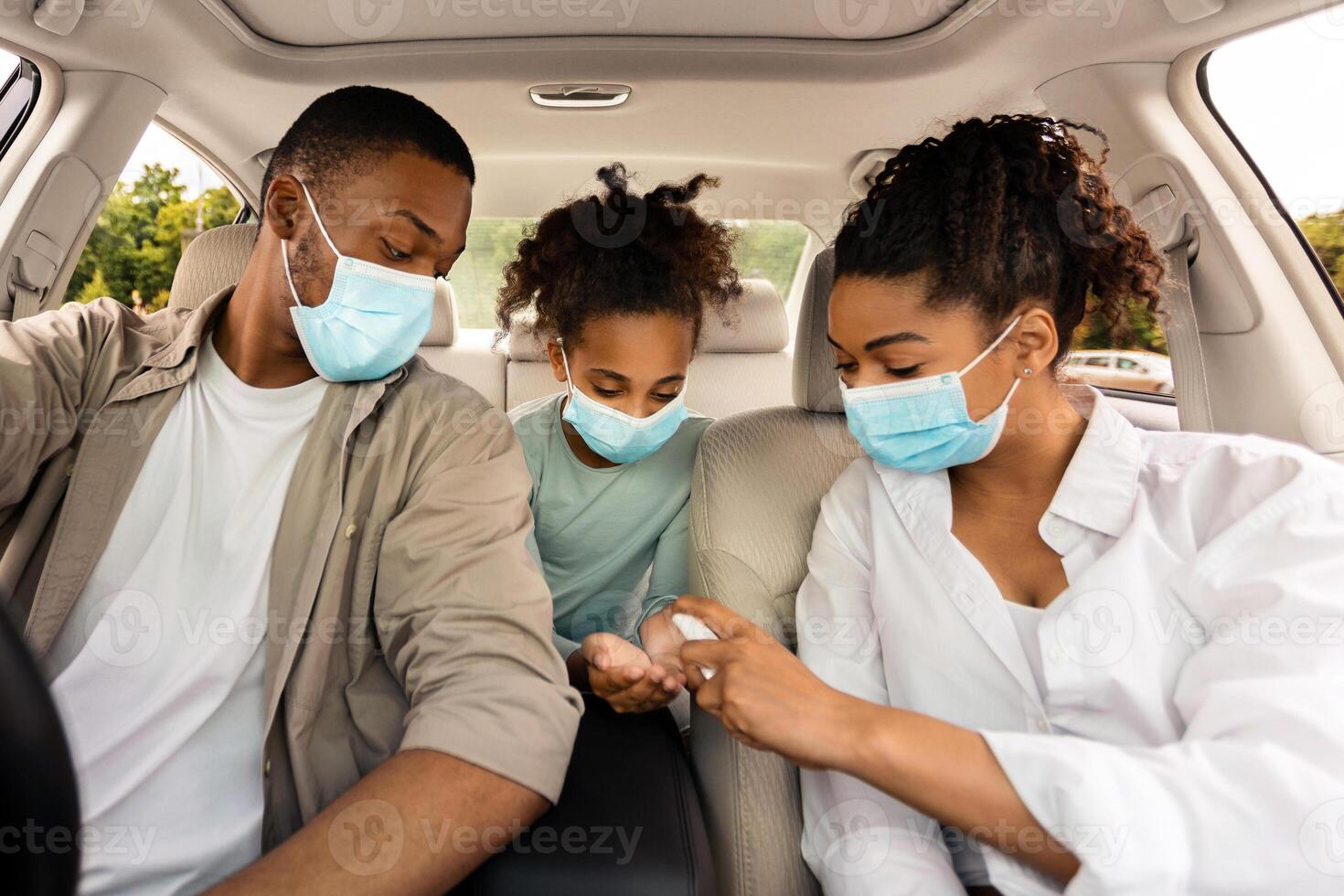 Black Family Spraying Sanitizer On Hands In Car. Parents And Daughter Traveling By Automobile, Wearing Face Masks And Disinfecting Hands For Covid-19 Protection photo