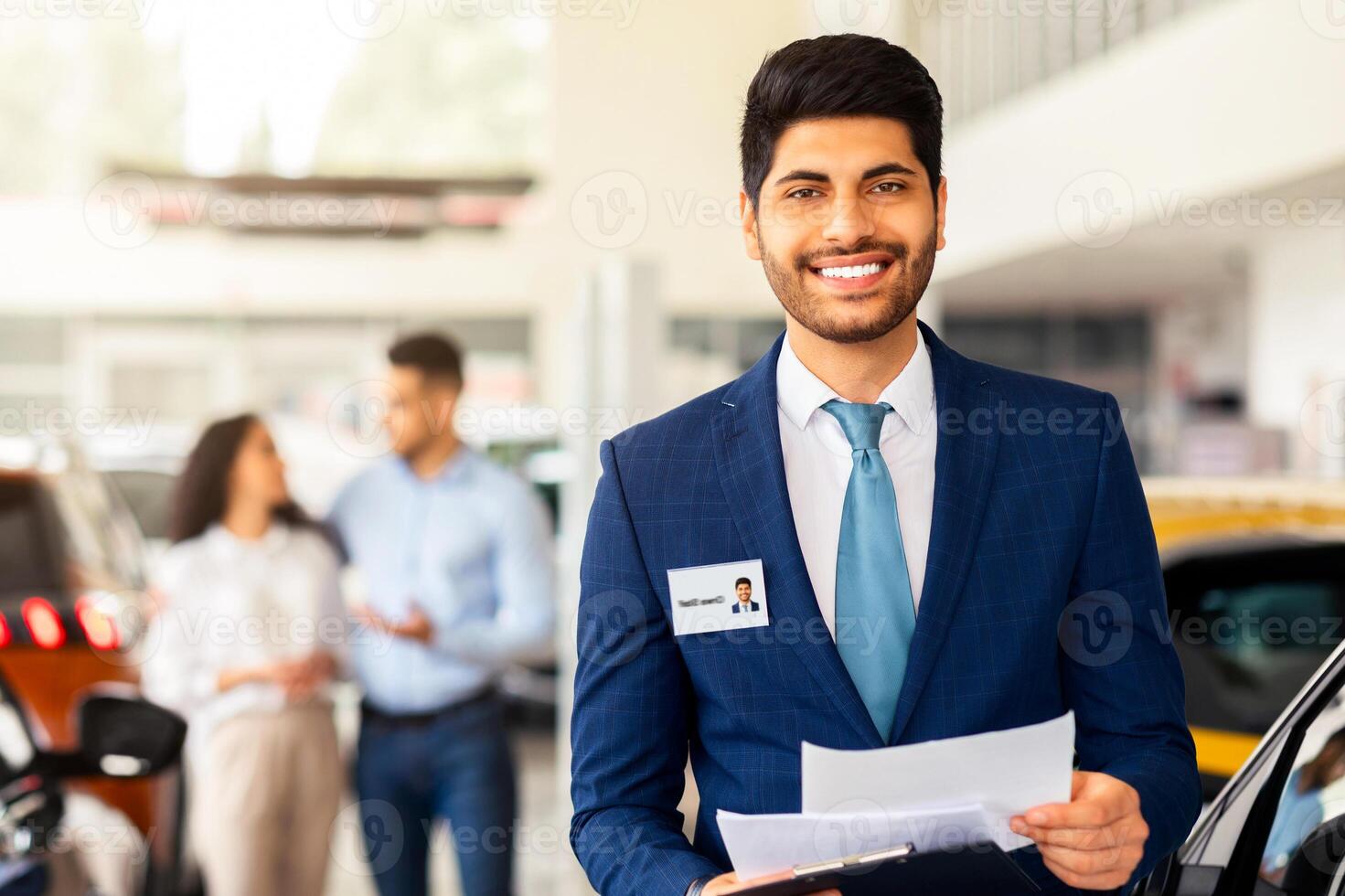 Charismatic arab man in suit sales assistant posing over auto showroom, holding chart with papers and smiling at camera, standing over various cars and customers choosing new automobile, copy space photo