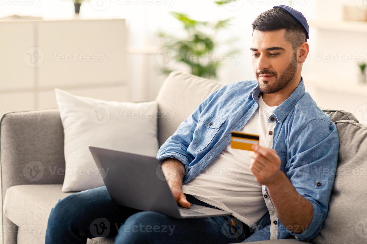 Electronic Payment Service Concept. Portrait of casual young jewish businessman in kippa holding credit card and using laptop, making e-payments while working with computer at home office, copy space photo