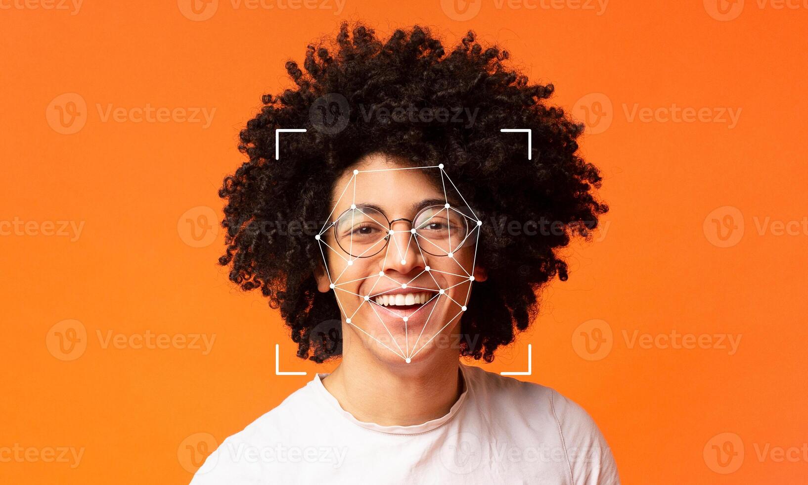 A person with curly hair and glasses smiles broadly, appearing cheerful. Their face is framed by a digital overlay indicating facial recognition technology against a vibrant orange backdrop. photo