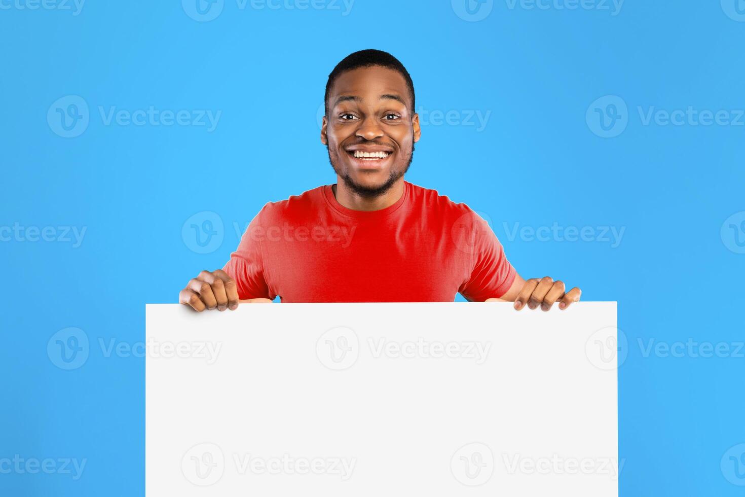 Look At This Offer. Black Guy Holding Empty White Paper Board Advertising Your Text Smiling To Camera Posing Over Blue Background. Studio Shot, Mockup. Sales And Discounts Advertisement photo