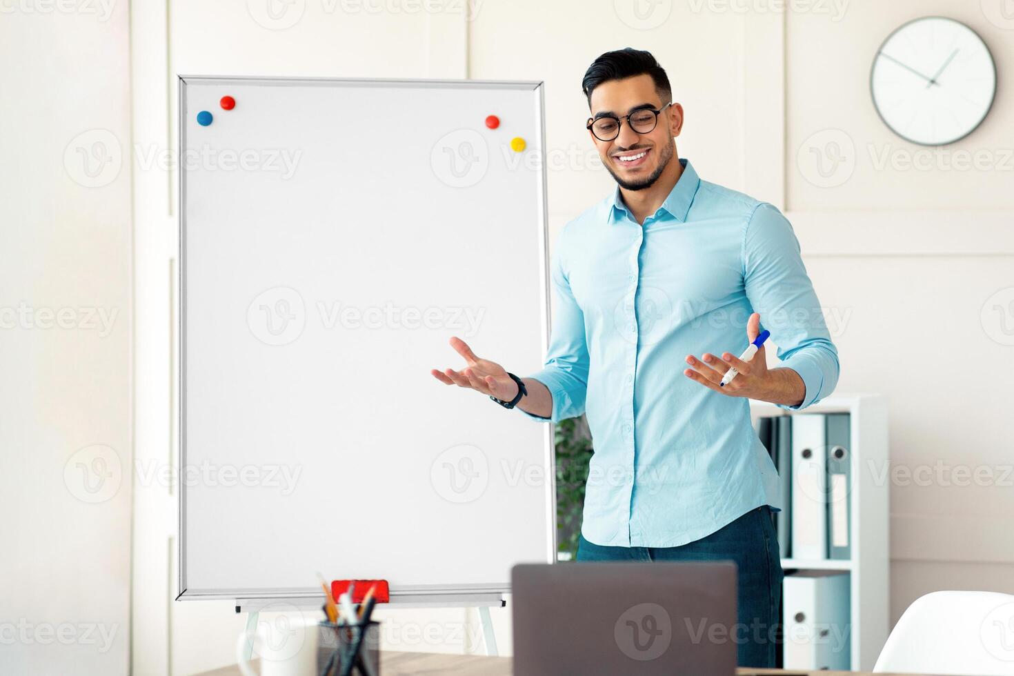 Cheerful young Arab teacher standing near blackboard and explaining English grammar to students online from home. Positive guy tutor explaining foreign language rules on web photo