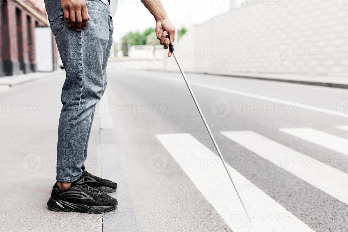 Cropped view of young black guy with vision disability standing on roadside, using walking cane in city centre, copy space. Unrecognizable millennial blind man waiting to cross road in downtown area photo