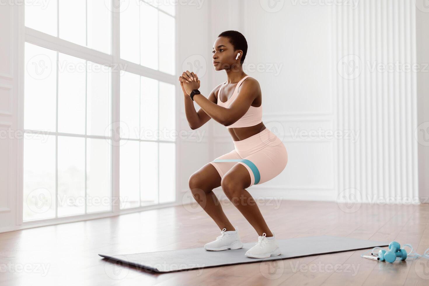 Body Shaping And Glutes Workout. Determined Fit Sporty Woman In White Sportswear Exercising With Resistance Loop Band Doing Squats Standing On Yoga Mat, Training Butt And Hamstring, Wearing Earbuds photo