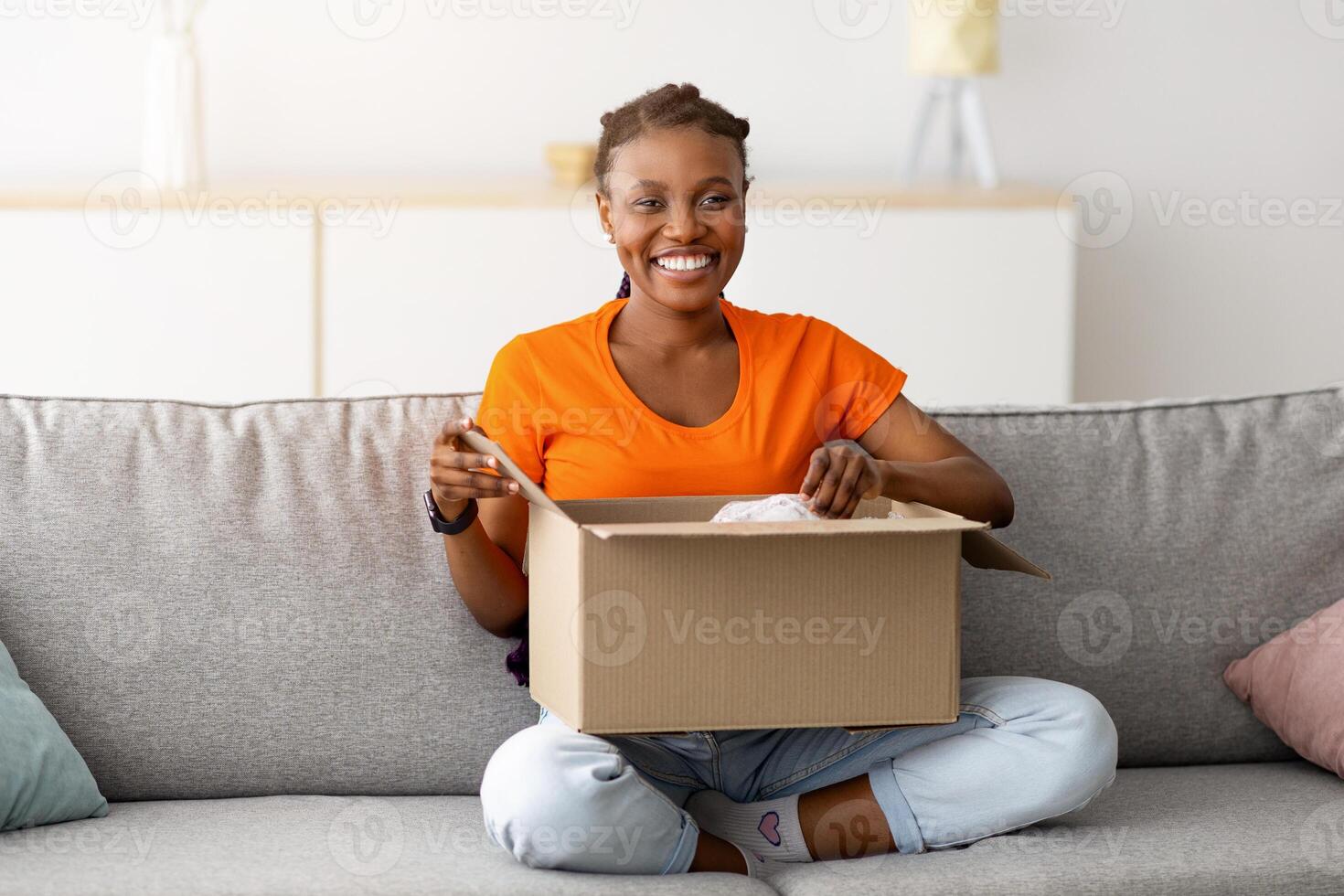 Full length of joyful black lady opening carton package, sitting on sofa with crossed legs, smiling at camera indoors. Young female consumer unboxing online store order, happy with delivery service photo