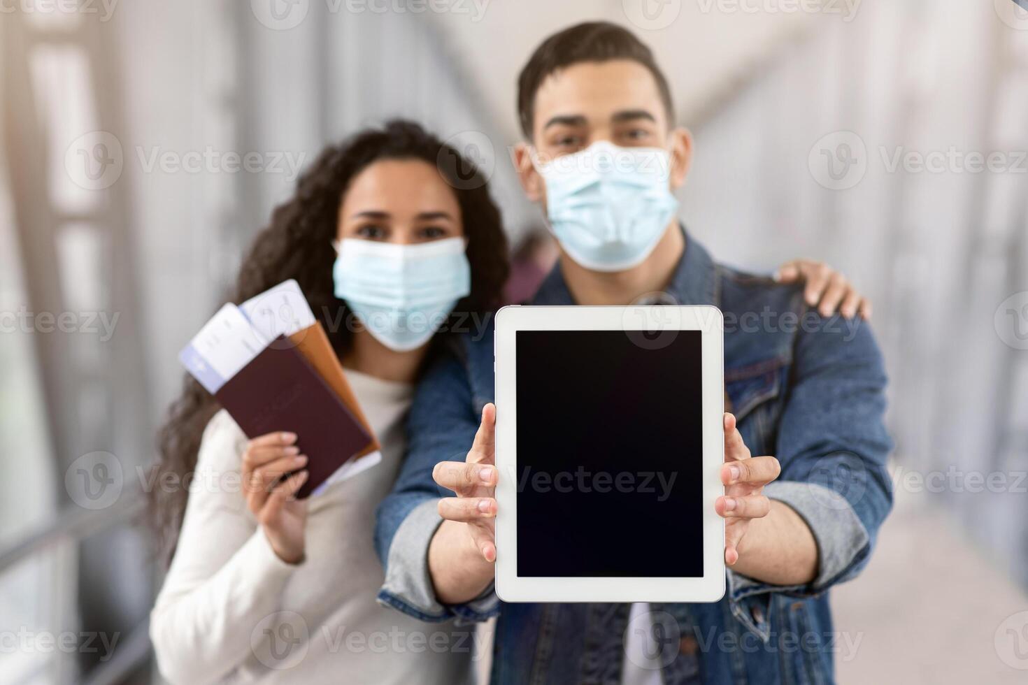 Arab Couple Wearing Medical Face Masks Demonstrating Digital Tablet With Black Blank Screen While Standing In Airport With Passports And Tickets, Spouses Showing Copy Space, Selective Focus, Mockup photo