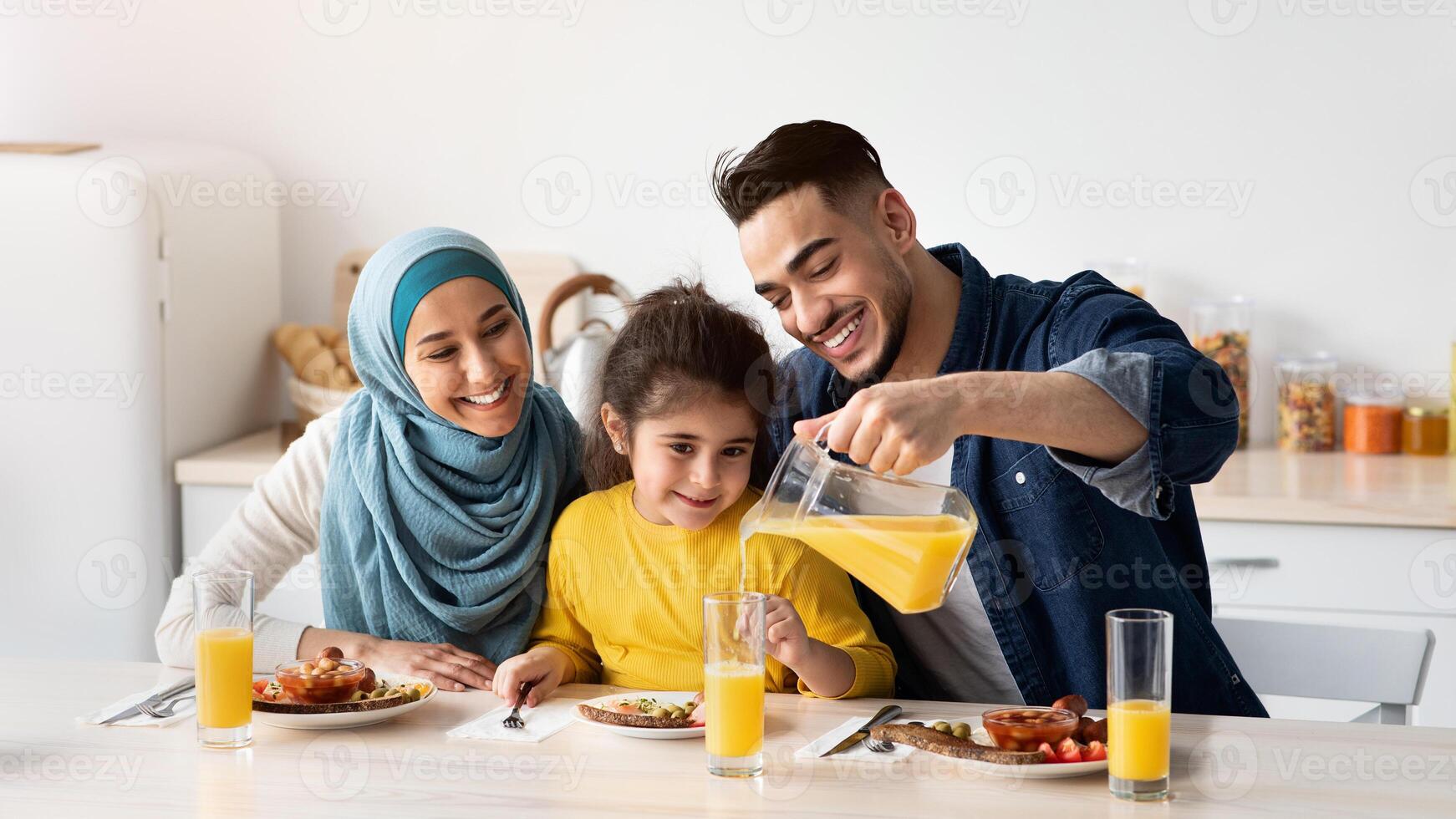 Portrait Of Happy Middle Eastern Family Of Three Having Breakfast In Kitchen Together, Arab Parents With Cute Little Daughter Enjoying Tasty Food And Drinking Orange Juice While Sitting At Table photo