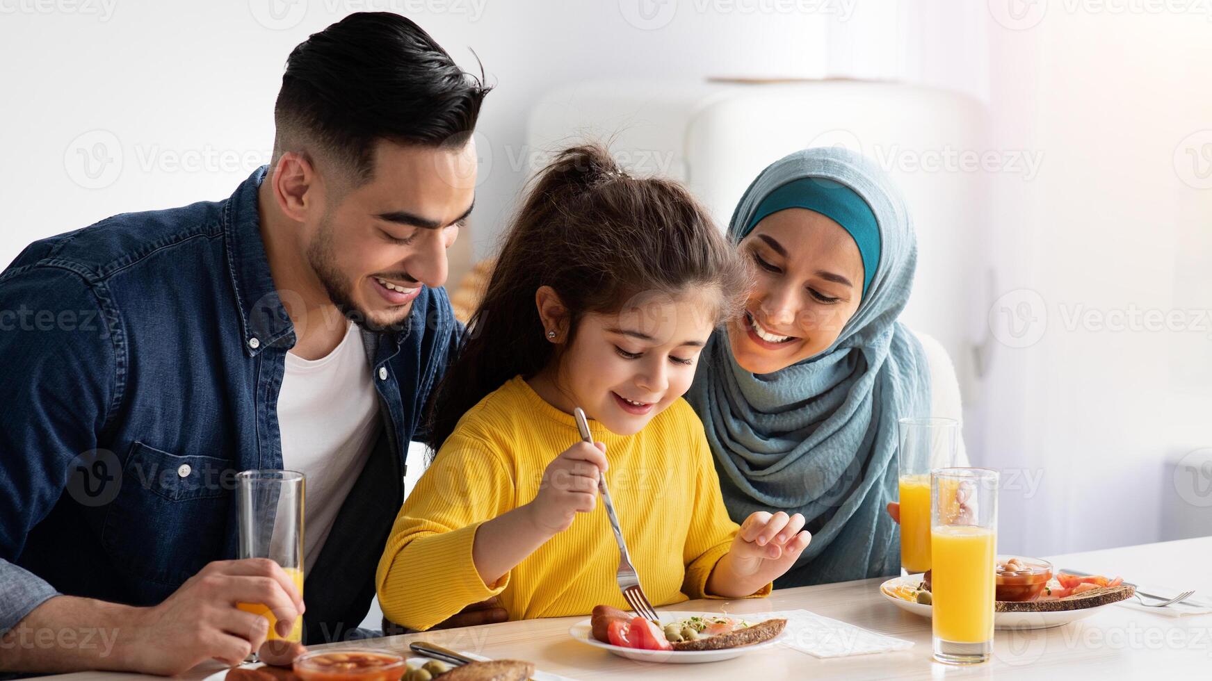 Tasty Breakfast. Happy Islamic Parents And Little Daughter Eating Together In Kitchen, Adorable Female Child Using Fork While Tasting Food, Happy Mom And Dad Looking At Their Kid, Closeup Shot photo