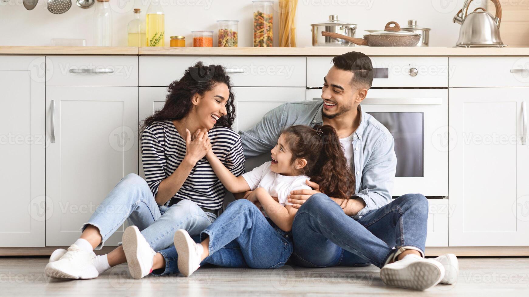 Beautiful middle eastern family father, mother and daughter sitting on floor at cozy white kitchen, loving parents enjoying time with their cute little child, having conversation, laughing, copy space photo