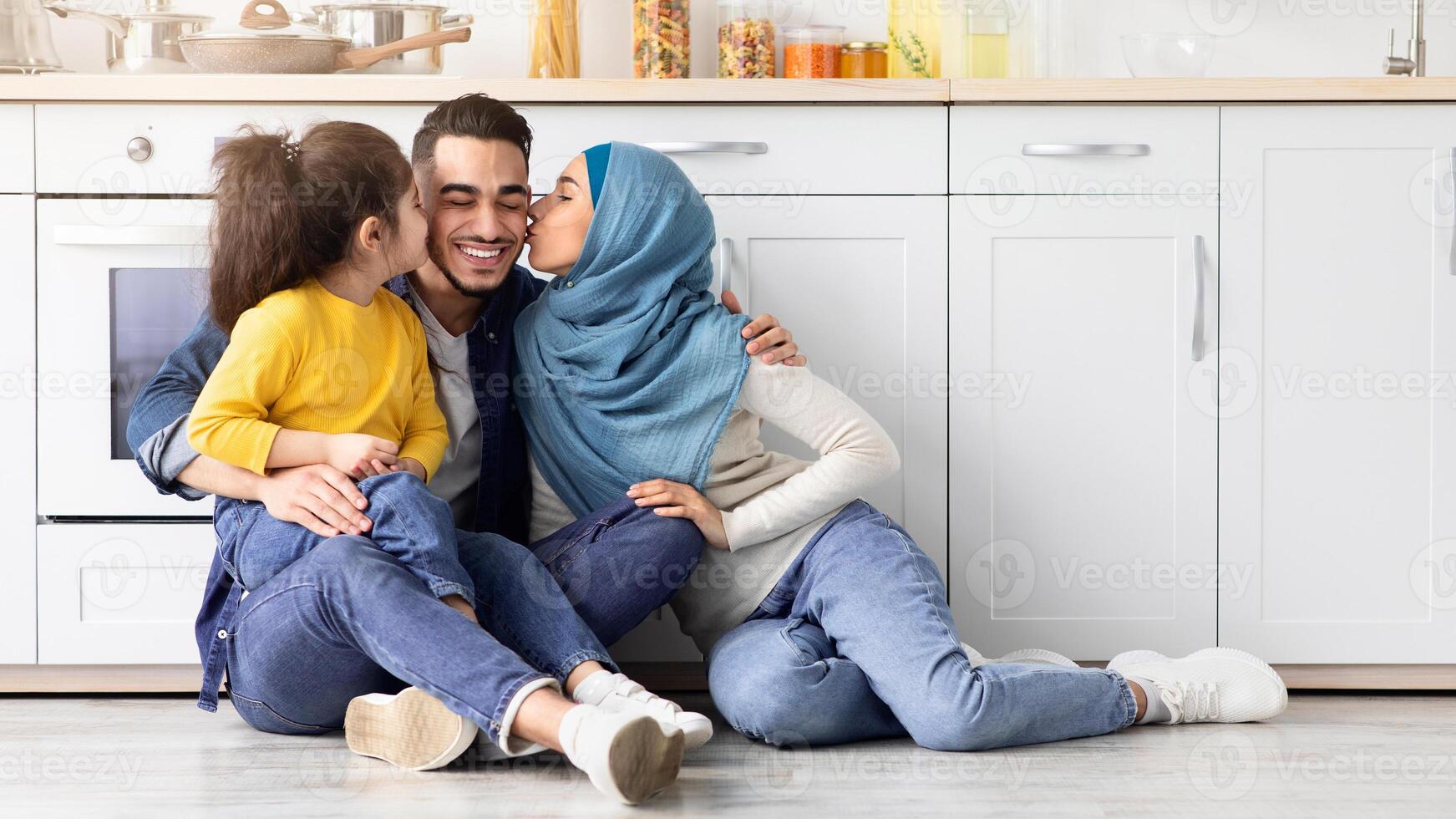 Portrait Of Loving Middle Eastern Family Of Three Spending Time Together In Kitchen, Cheerful Muslim Mother And Little Daughter Sitting On Floor And Kissing Happy Father To Cheeks, Free Space photo