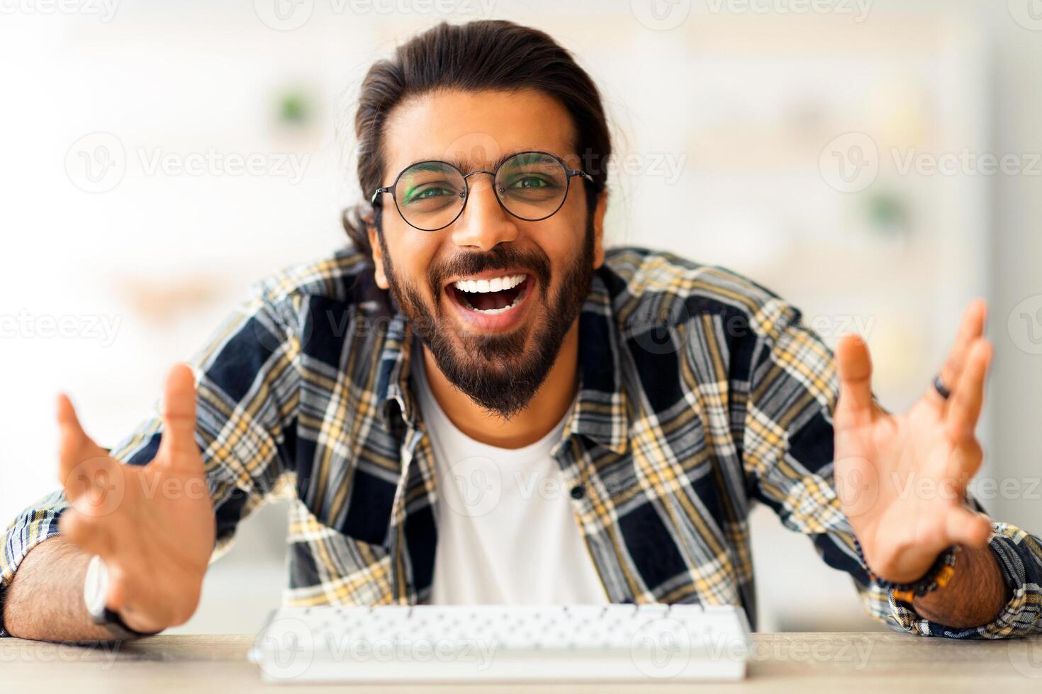 Emotional middle-eastern guy freelencer having conference with someone, sitting at worktable with computer keyboard, smiling at pc camera and cheerfully gesturing, home interior, copy space photo