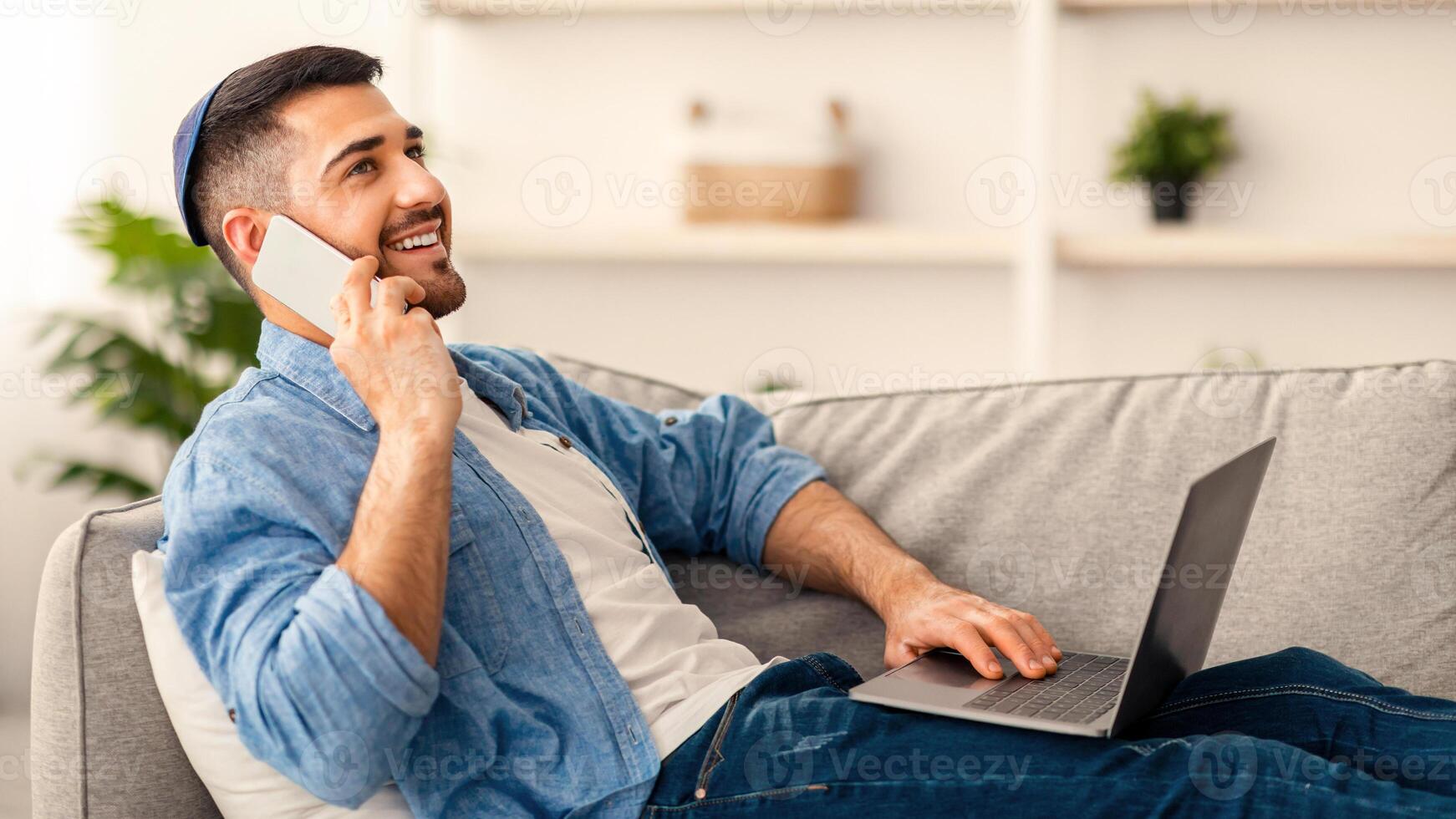 Distant Work and Telecommunication. Happy male jewish freelancer talking on cellphone and using laptop working remotely from home, sitting on sofa in living room, free space, selective focus photo