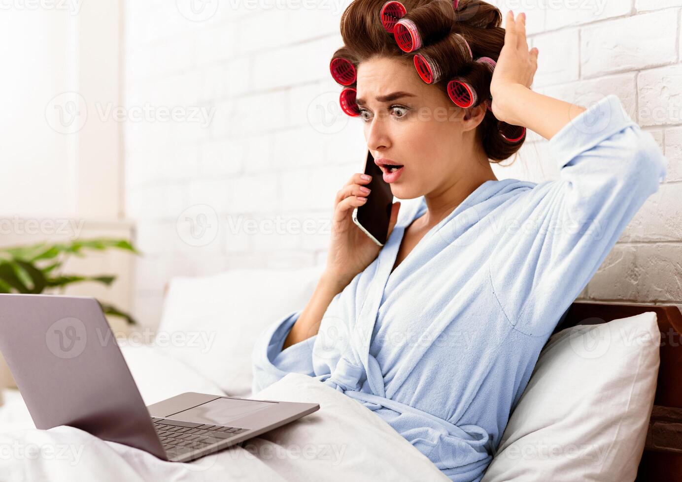 Shocked Housewife Talking On Phone Using Laptop Browsing Internet Lying In Bed At Home. Shocking News Concept photo