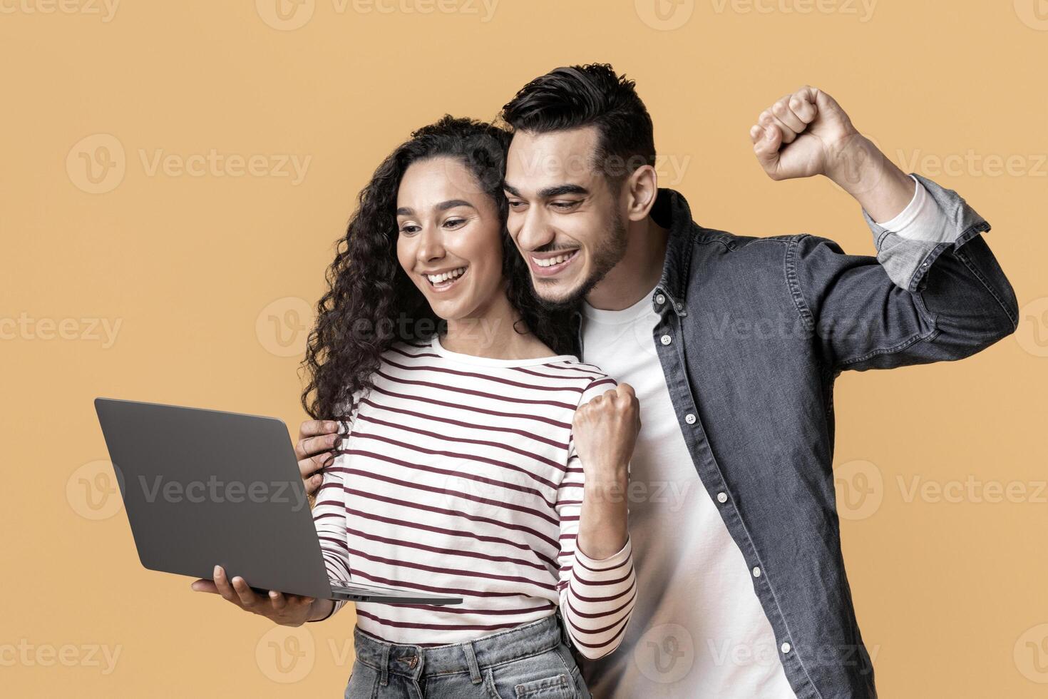 Online Win. Joyful Middle Eastern Couple Holding Laptop And Celebrating Success, Happy Young Arab Man And Woman Looking At Computer Screen And Shaking Fists, Standing On Yellow Background, Copy Space photo