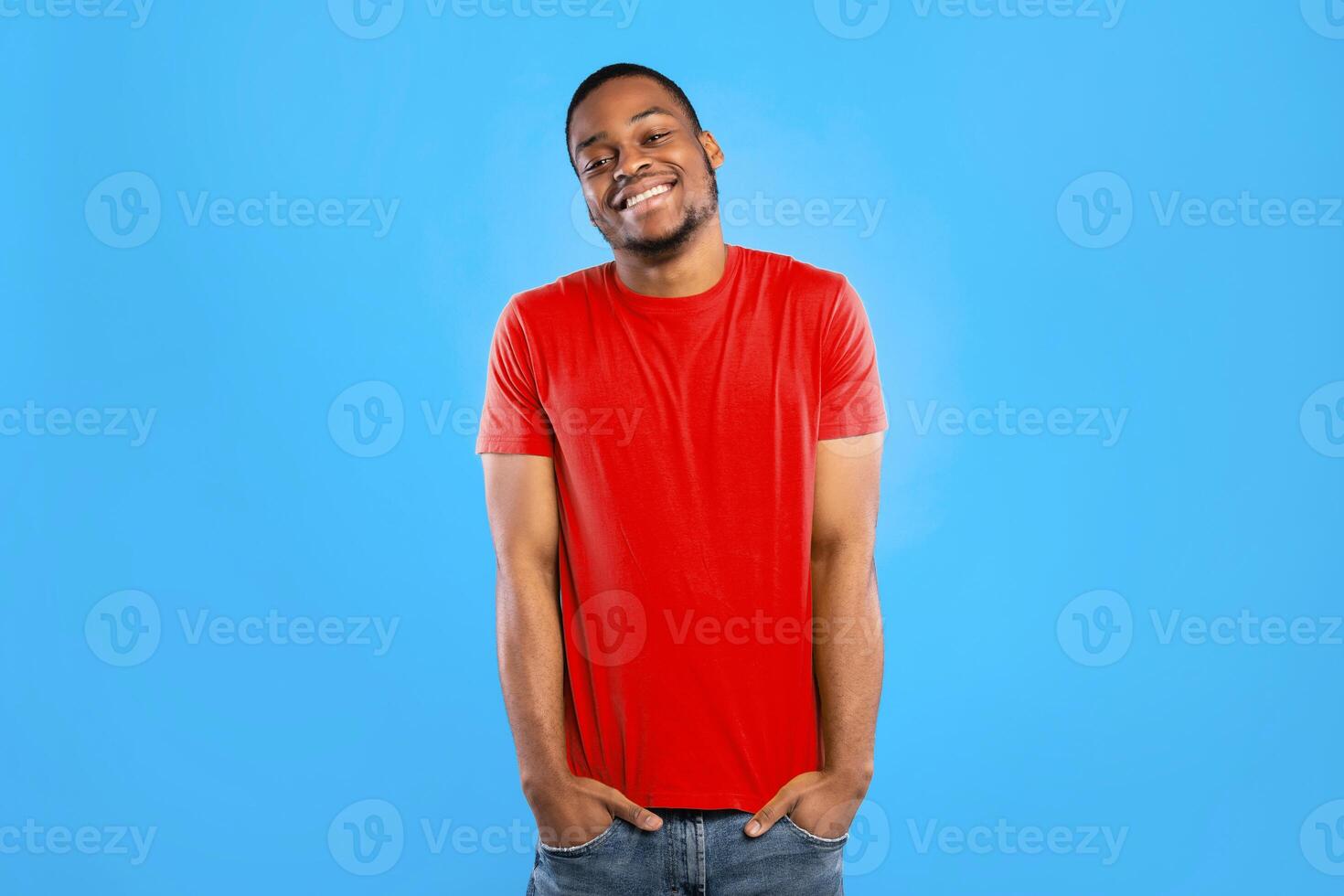 Happy African American Man Wearing Bright Yellow T-Shirt Standing Holding Hands In Pockets Posing Over Blue Background In Studio. Shot Of Successful Millennial Guy Smiling To Camera photo