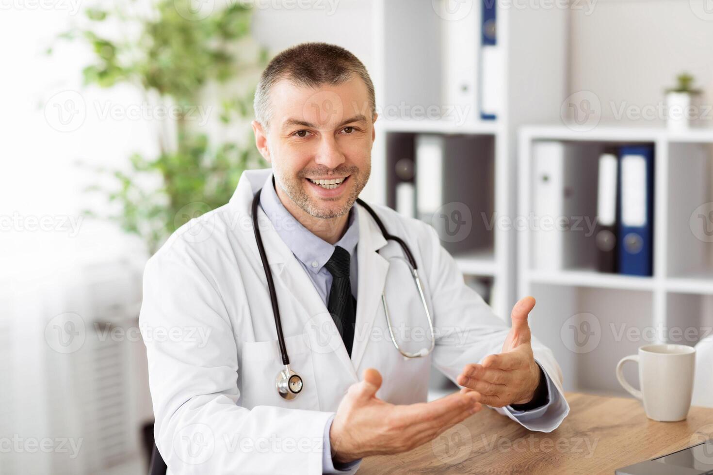 Distance Therapy. Portrait of mature male doctor talking online with patient, making call, looking at camera, adult specialist wearing white uniform with stethoscope speaking, consulting photo