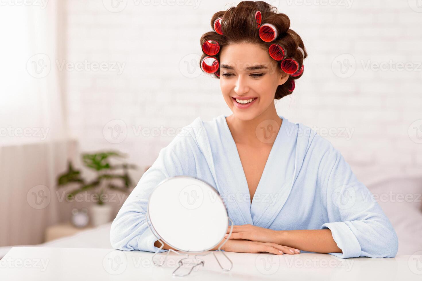 Hairstyling. Charming Lady Curling Hair With Curlers Sitting In Front Of Round Mirror In Modern Bedroom At Home. Female Beauty Routine Concept. Selective Focus, Empty Space For Text photo