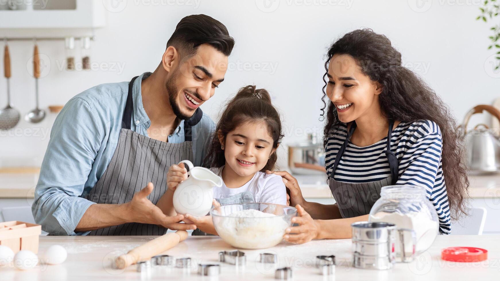 Happy middle-eastern family making homemade cookies together at kitchen, panorama. Joyful arab father, mother and little girl daughter pouring milk into bowl with dough, making pastry at home photo