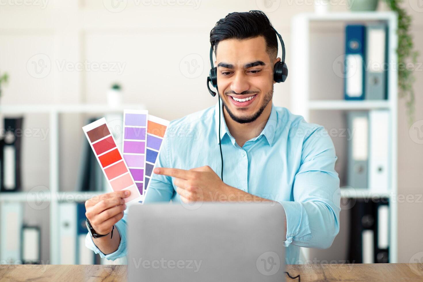 Young Arab man holding color swatches in front of laptop at office, presenting graphic design project during online company meeting. Millennial designer selecting gamma for future home interior photo