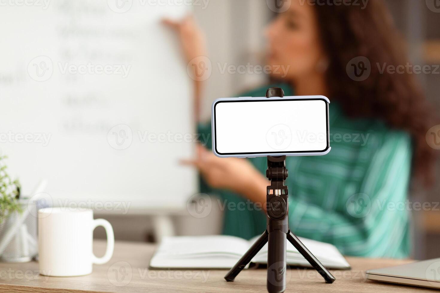 Modern Remote Education. Lady teacher shooting lecture on smartphone using tripod having conference with students, explaining material. Selective focus on blank white gadget screen, mock up photo