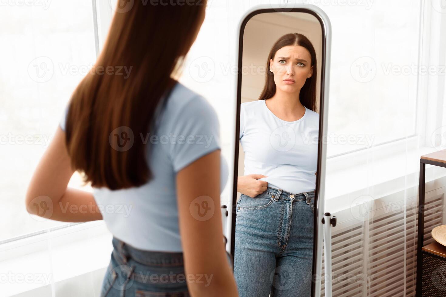 Weight Gain. Frustrated Girl Touching Belly Struggling To Lose Excess Kilograms Standing At Mirror Slimming At Home. Selective Focus photo