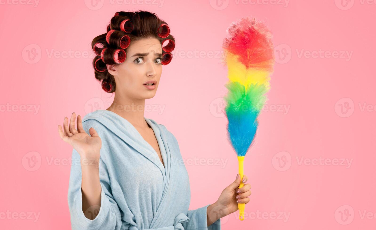 Spring Cleaning. Confused Glamorous Lady With Hair Curlers Holding Feather Duster Doing House Chores Looking At Camera In Shock Standing Over Yellow Studio Background. Housework Concept photo