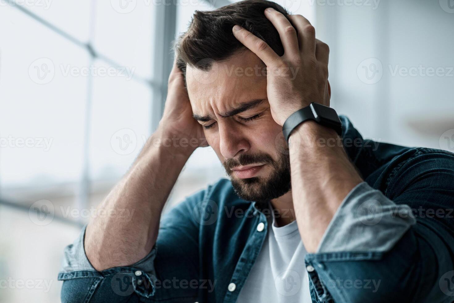Stress, headache, health problems, negative emotions. Despair sad unhappy young european handsome man holding his head, suffering from pain, overwork on large window background and crying, close up photo