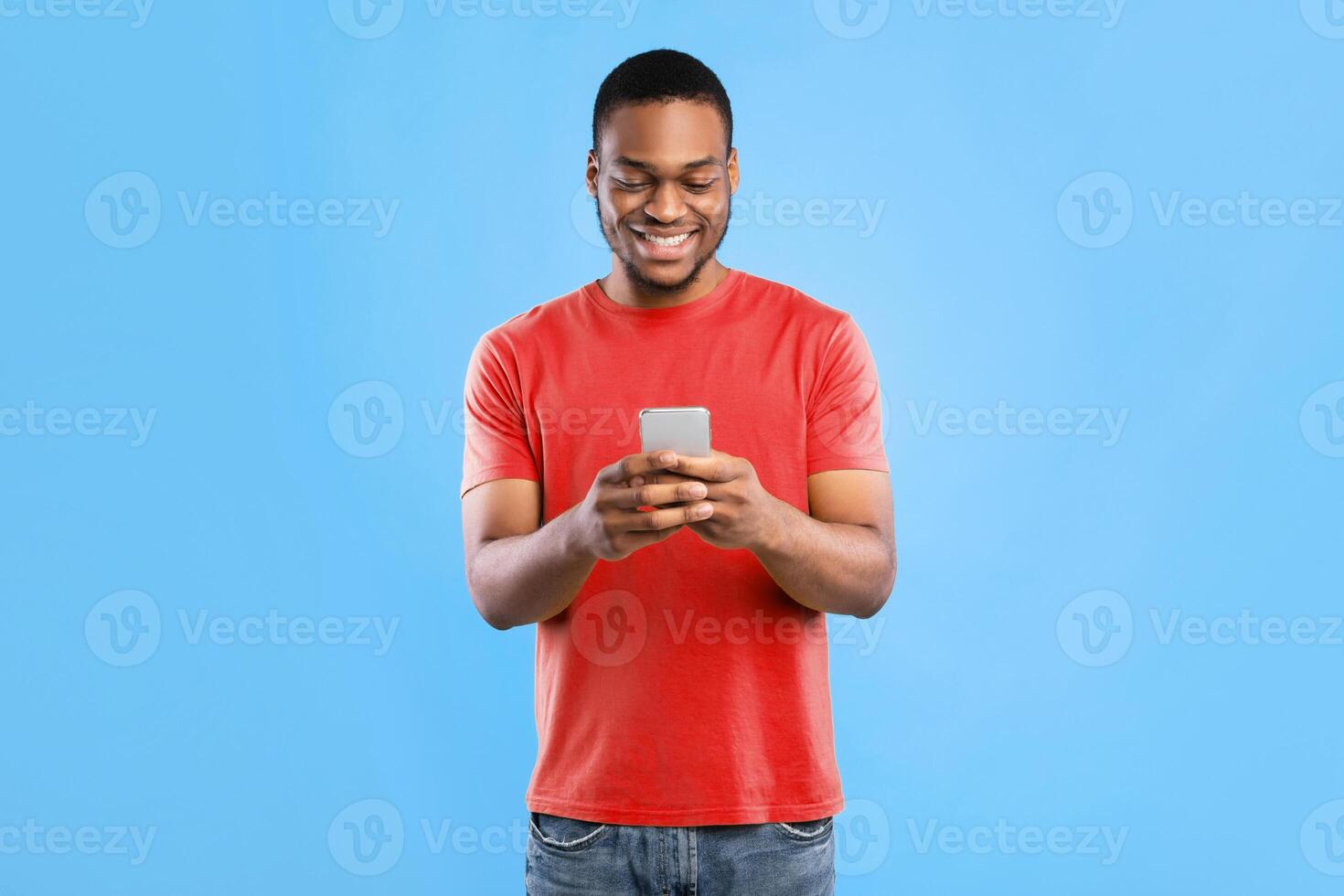 Happy African American Male Using Mobile Phone Texting Standing Over Blue Studio Background. Cheerful Black Guy Browsing Internet. New Application For Your Smartphone. People And Gadgets photo