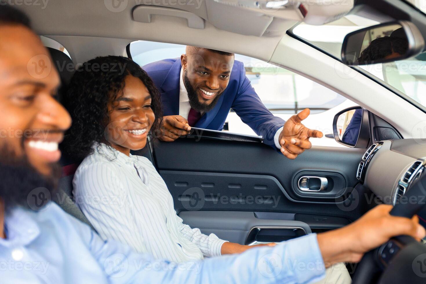 Handsome Salesman Presenting New Automobile To Young Black Couple In Showroom, African American Spouses Sitting Inside Of Car And Testing Vehicle Characteristics Before Buying In Dealership Center photo
