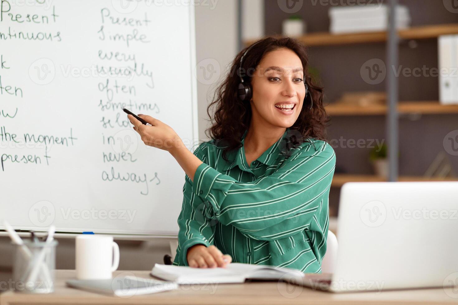 Modern Distance Learning. Smiling English teacher making conference chat with students using pc laptop. Lady sitting at desk pointing on board teaching from home during covid outbreak photo