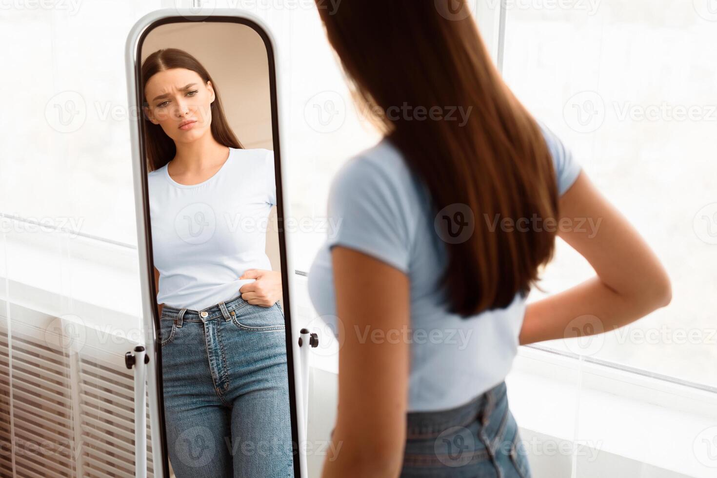 Weight Gain. Unhappy Girl Touching Drooping Belly Standing In Front Of Mirror At Home. Empty Space, Selective Focus photo