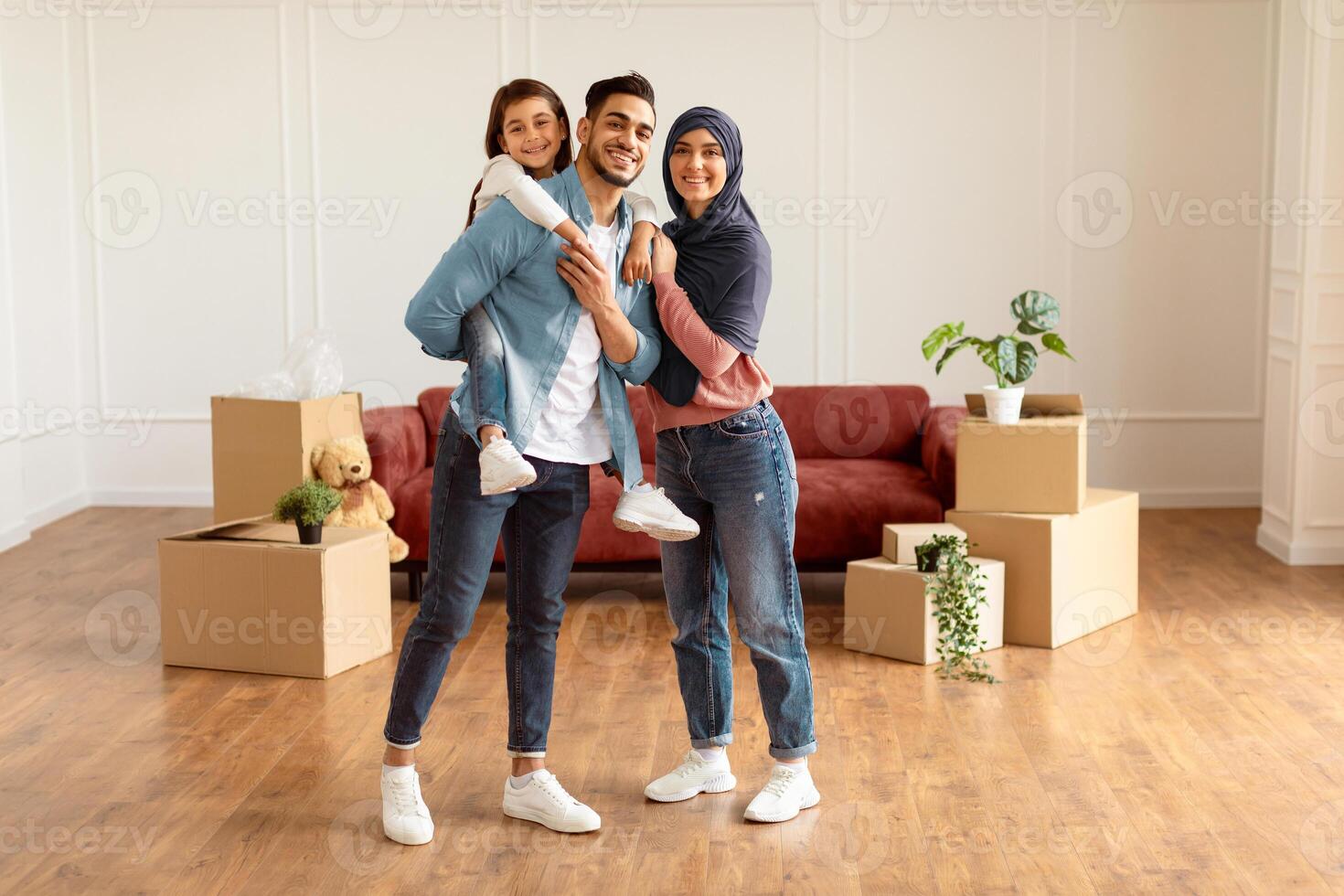 Full body length of happy islamic dad, mum in headscarf and little girl posing in new apartment, standing in empty living room with cardboard boxes. Cheerful father carrying his daughter on back photo