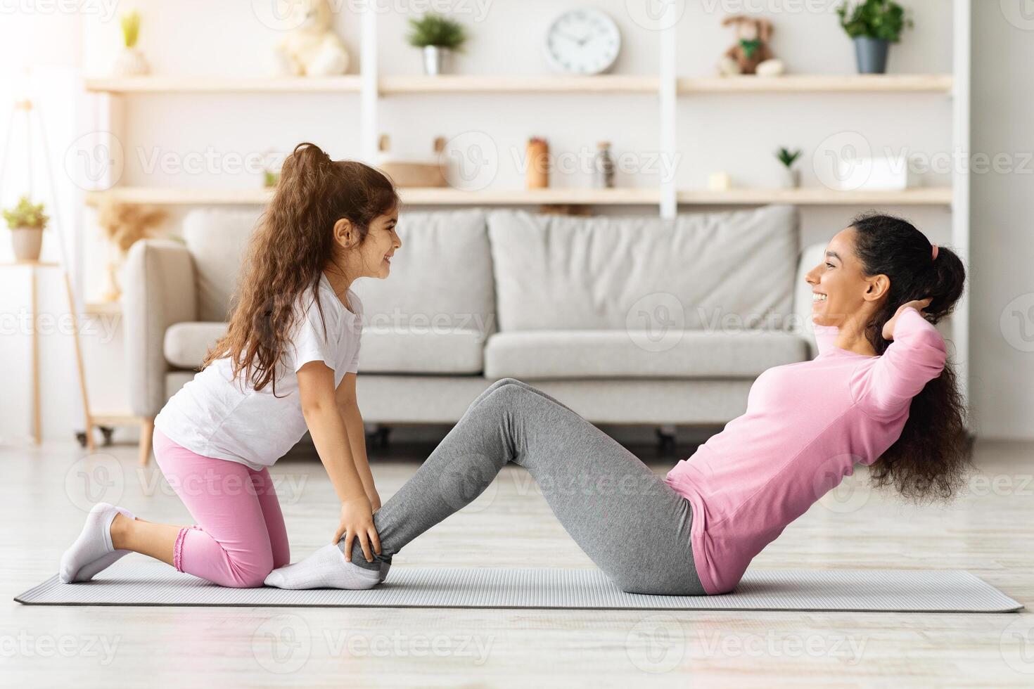 Cute mother and daughter working on abs muscles together at home, adorable little girl holding her mom legs while she doing crunches, exercising together in living room, side view, copy space photo