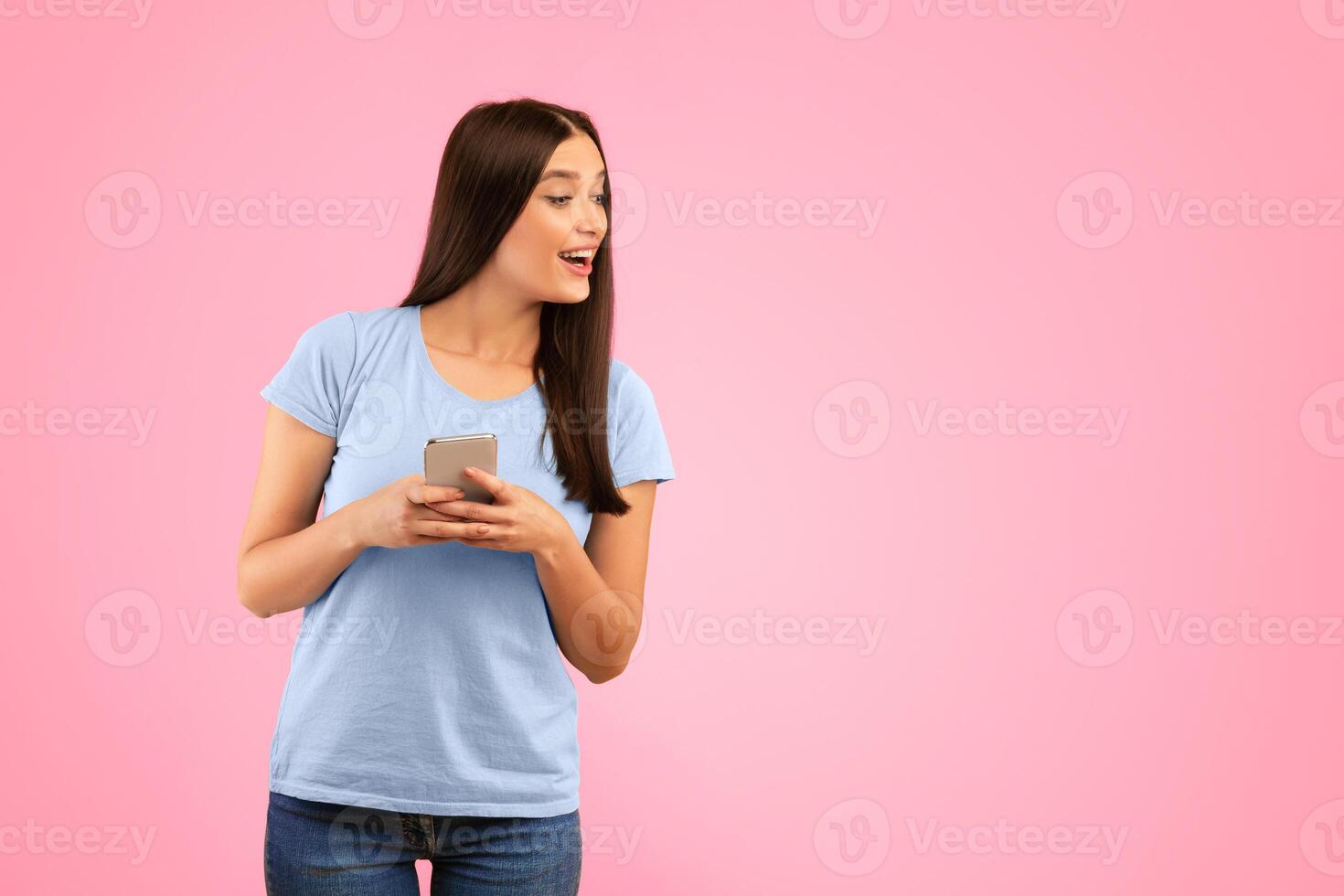 Portrait of smiling curious woman using smartphone standing isolated over yellow studio background, looking away, peeking and trying to see something, holding gadget, copy space. Lady chatting online photo