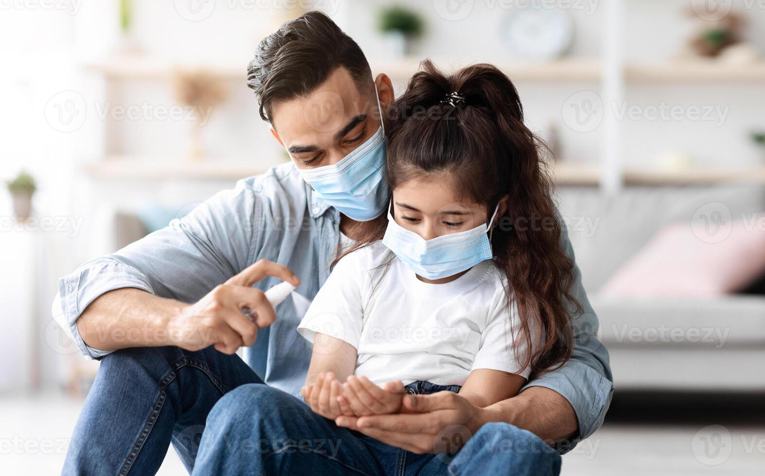 Middle-eastern father and daughter in protective face masks sitting on floor in living room, using disinfectant after coming back home from the street. Family protection while coronavirus pandemic photo