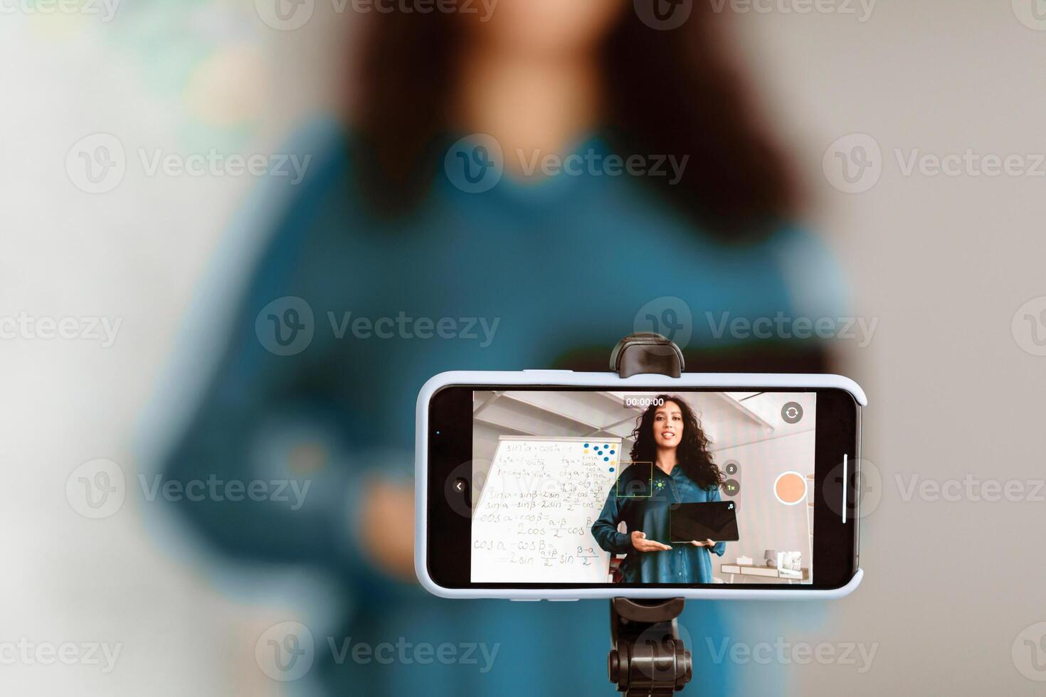 Closeup of smart mobile phone taking live of lady teacher giving lesson with math formulas on black board in school classroom, holding showing tablet, technology live streaming and broadcast concept photo