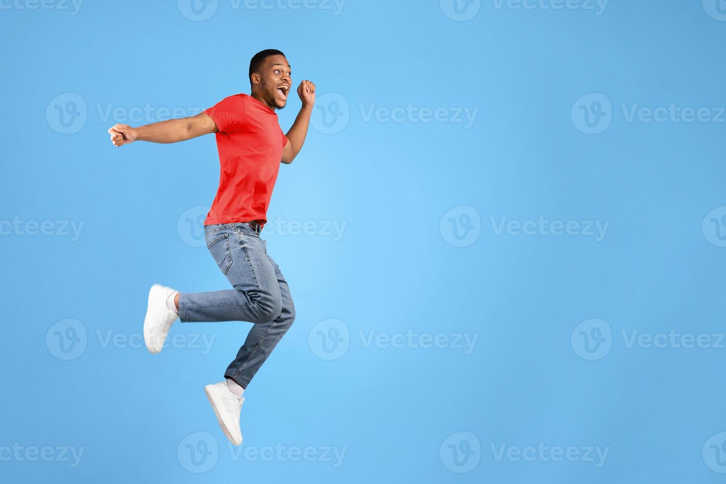 Joyful African American Millennial Man Jumping And Shouting In Joy Posing In Mid-Air Over Blue Studio Background. Positive Emotions And Success Celebration Concept photo