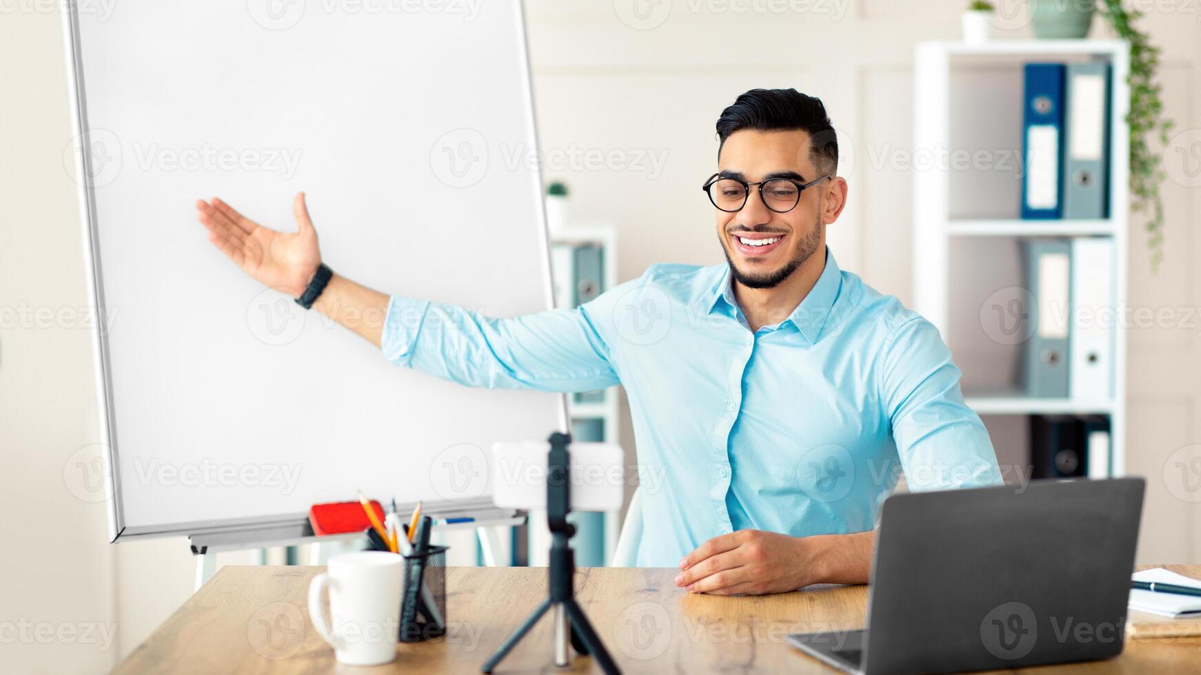 E learning concept. Positive Arab guy teacher giving online English lesson on smartphone, pointing at blackboard with grammar rules at home. Young tutor having web conference with students photo