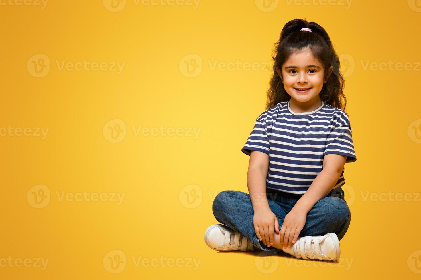 Portrait Of Adorable Little Middle Eastern Girl Posing Over Yellow Background, Cute Small Arab Female Child In Casual Clothes Sitting With Legs Crossed And Smiling At Camera, Copy Space photo