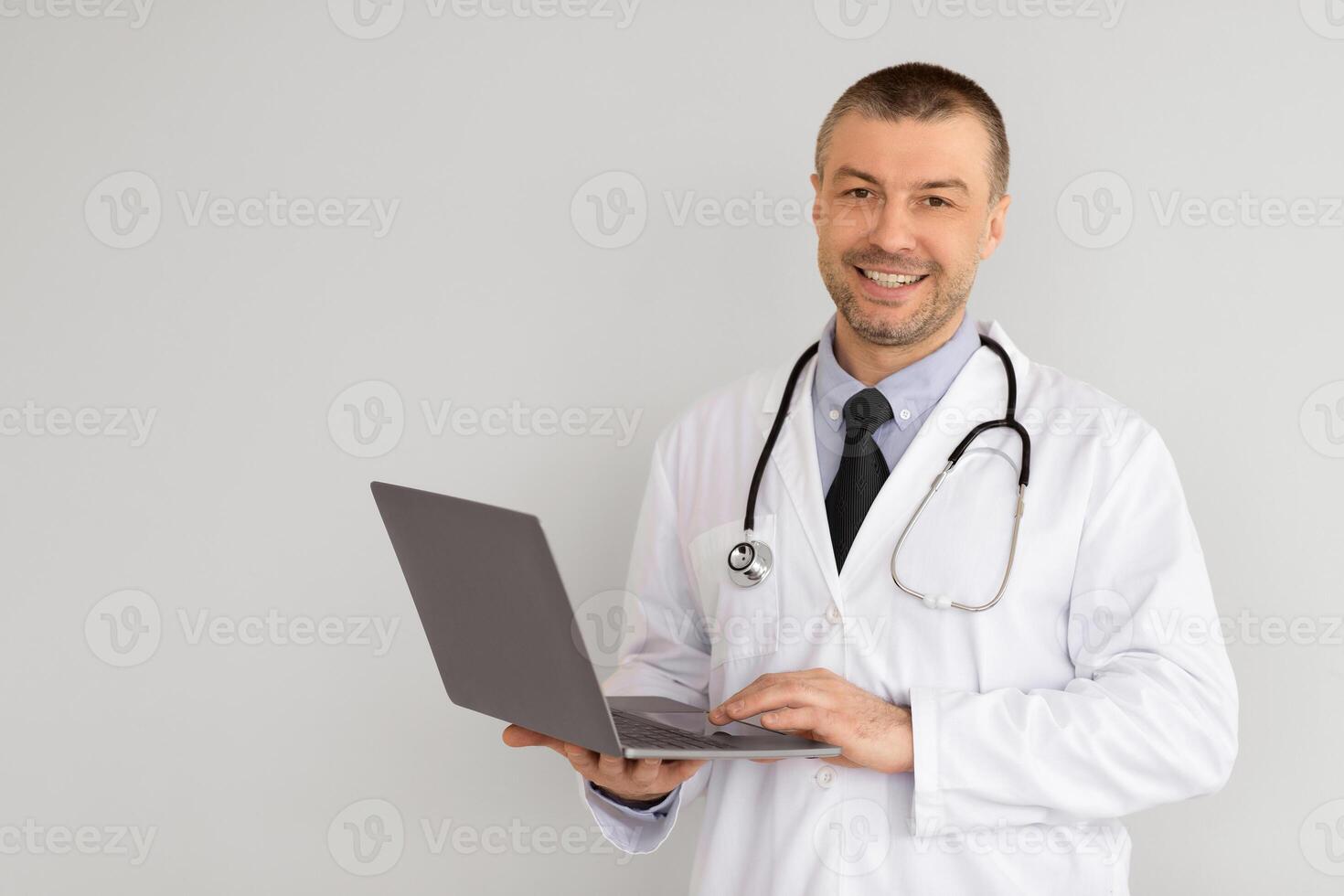 Health Care And Telemedicine Concept. Portrait Of Smiling Confident General Practitioner Wearing Coat And Stethoscope Holding And Using Laptop, Standing Isolated On White Studio Background, Copy Space photo
