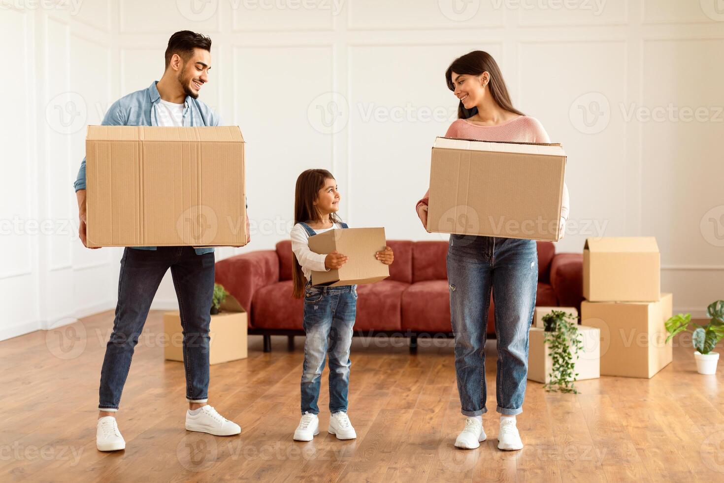 Full Body Length Portrait Of Cheerful Family Of Three People Housing, Happy Smiling Man, Woman And Little Girl Holding And Carrying Cardboard Boxes Standing In Empty Living Room. House Move Concept photo