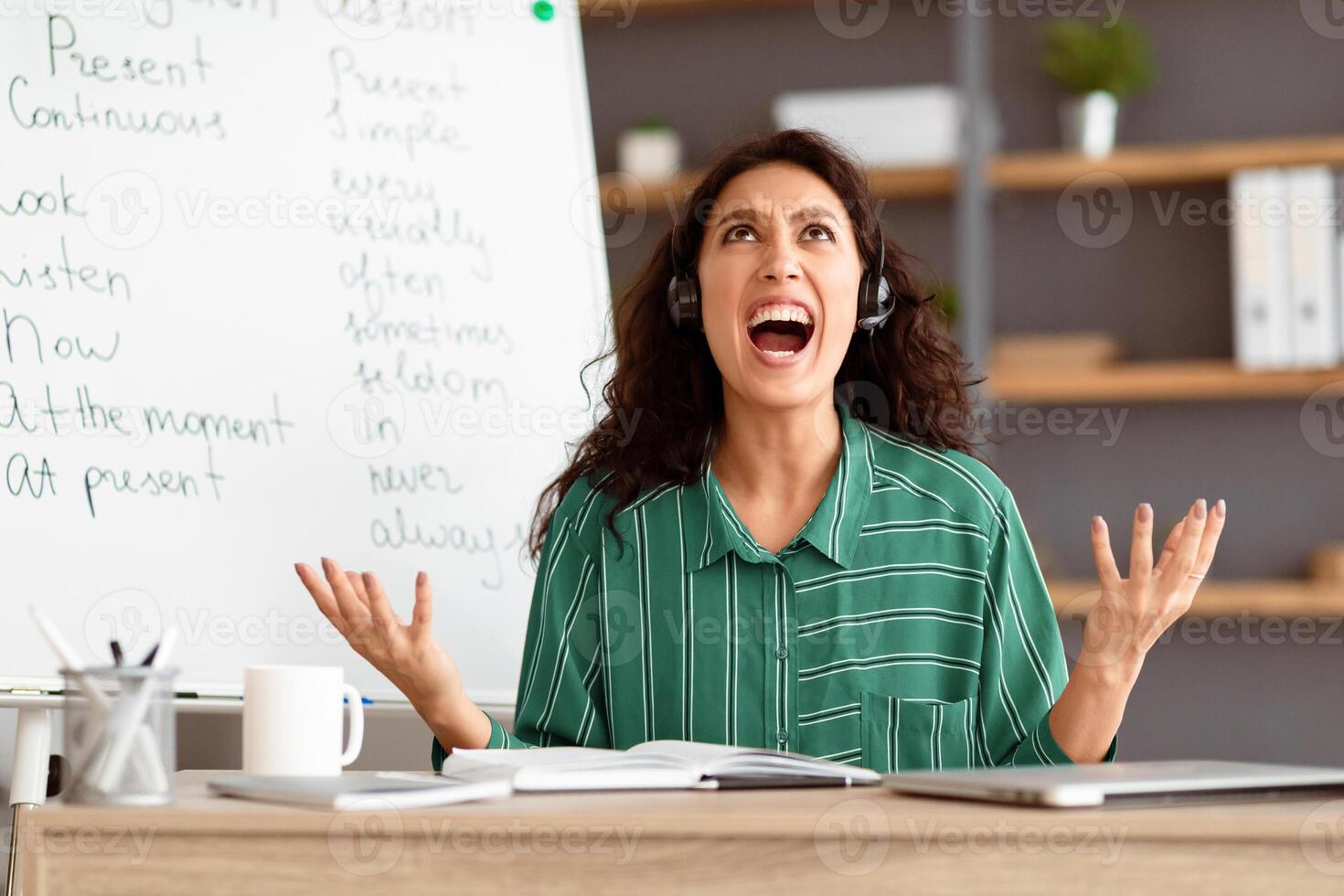 Fatigued From Work. Tired young teacher in headset screaming out loud, gesturing and looking up, having lesson with students, tutoring English language online, sitting at desk and yelling photo