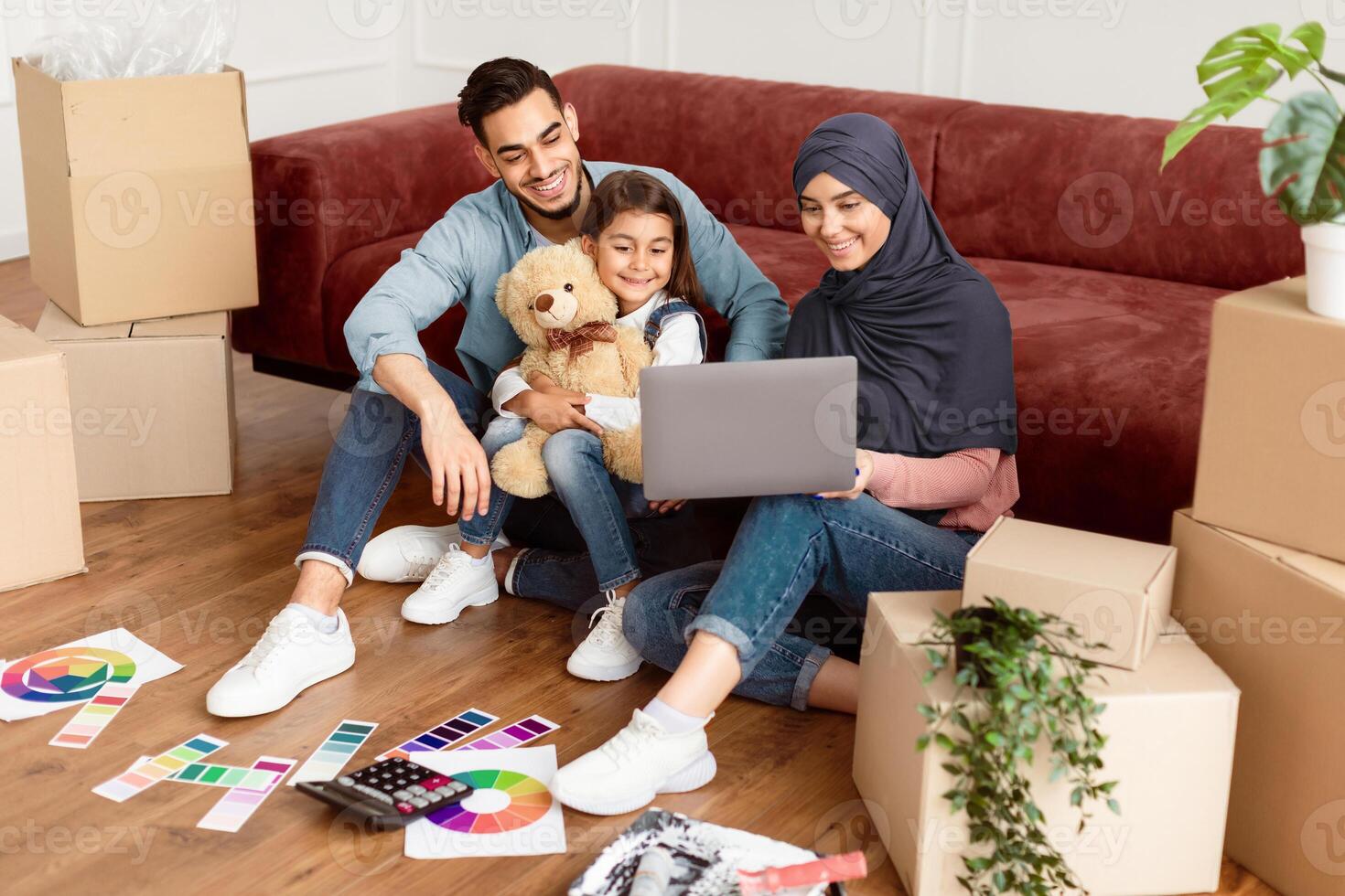 Relocation Concept. Joyful Middle Eastern Family Of Three People Using Laptop Sitting On Floor Among Moving Boxes And Paint In New House, Choosing And Ordering Furniture, Discussing Decor And Design photo